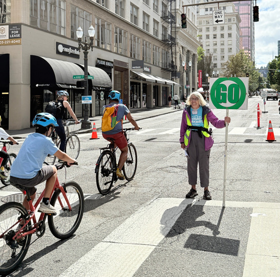 A smiling volunteer holds an event sign as Sunday Parkways participants enjoy Downtown Portland