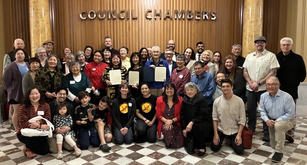 Large group of people in rows, standing in front of wooden wall with "Council Chambers" sign. Some are holding mayoral proclamations.