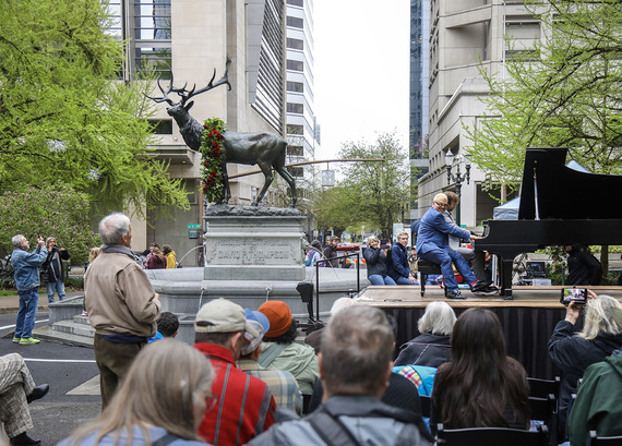 Large bronze elk statue tops a granite pedestal and fountain, adorned with a wreath of red roses while two pianists perform