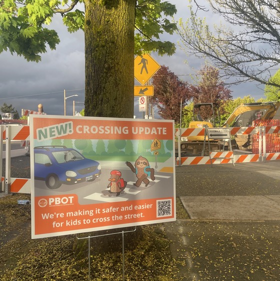 An orange yard sign announcing a new crossing update sits in front on active construction near a sidewalk.