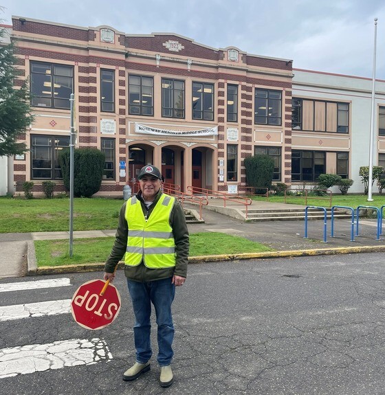 An adult wears a high-visibility traffic safety vest and holds a stop sign while standing in front of a school.