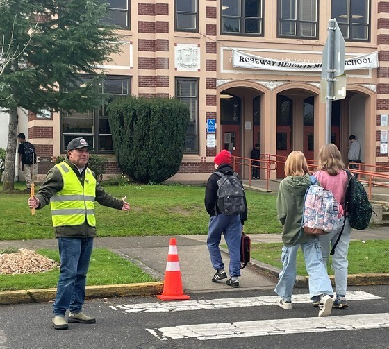 An adult wears a high-visibility traffic safety vest and holds a stop sign while ushering students across a crosswalk.