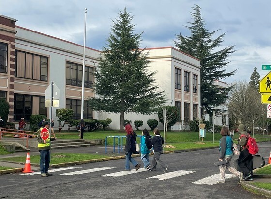 An adult wears a high-visibility traffic safety vest and holds a stop sign while ushering students across a crosswalk.