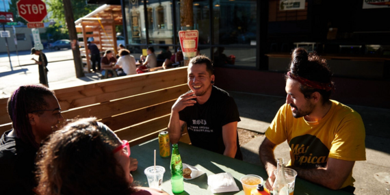 A diverse group of friends laugh at an outdoor dining table on a Portland street, closed for pedestrians