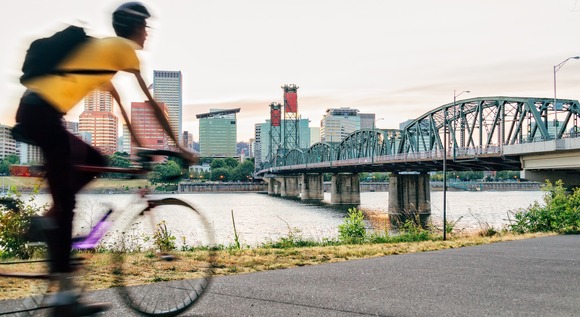 A cyclist rides the Eastbank Esplanade at sunset, with the Hawthorne Bridge, Willamette River, and downtown Portland glowing softly in the background
