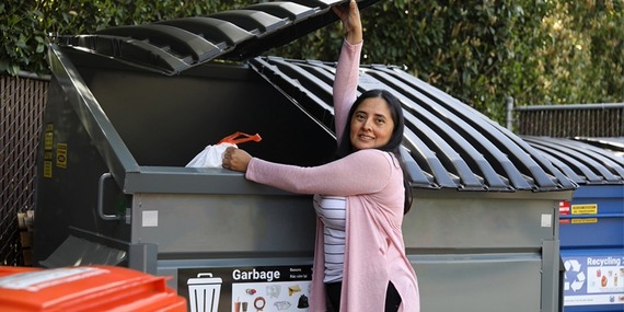 A woman smiles while opening a dumpster to throw out garbage and recycling
