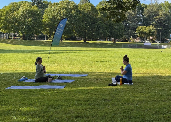 Two people are seated on mats at a city park, hands together and facing each other for a Fitness in the Park class