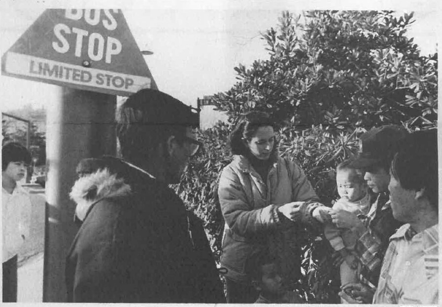 Black and white image of people gathered around a person speaking to them at a bus stop, circa 1981