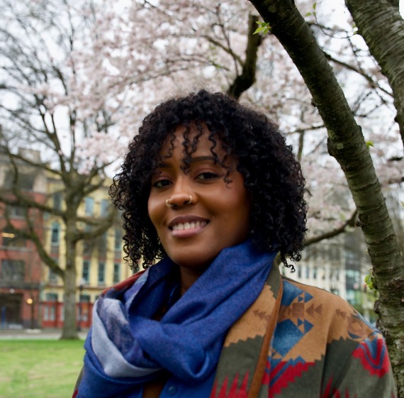 ARTS - Headshot of Hanifah Abioto standing in a natural area in front of buildings.