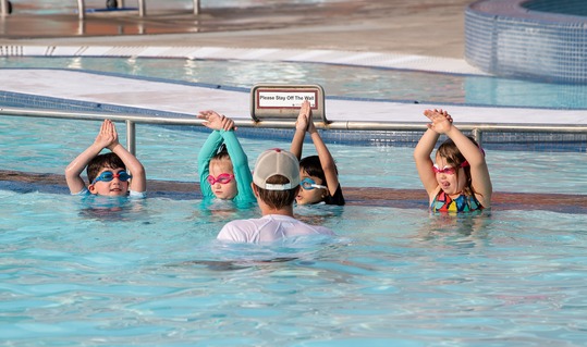 four kids  in a swim lesson at an outdoor pool stand in the water facing the instructor with their hands above their heads. 