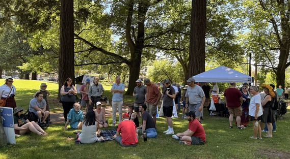 Large group of people standing and sitting on lawn under large trees, with white tent in background