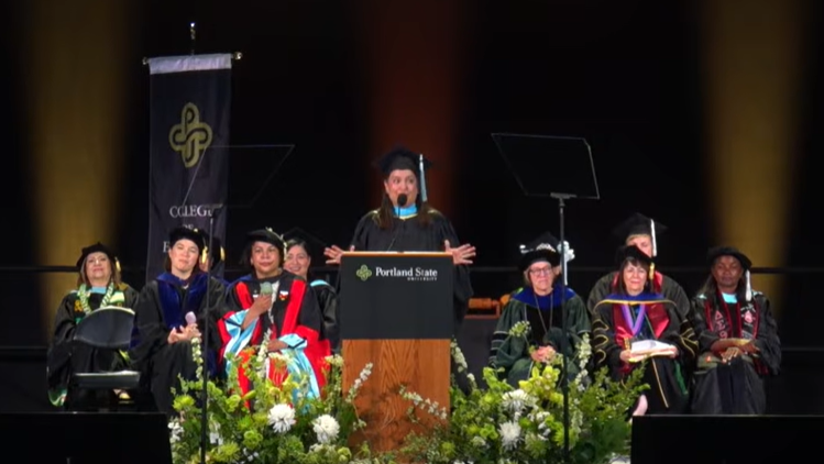 Councilor Koyama Lane in cap and gown at Portland State University lectern, in front of line of seated people in academic regalia