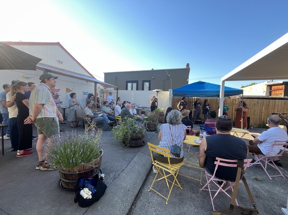 Gathering of people, some standing and some sitting, watching a speaker under a blue pop up tent 