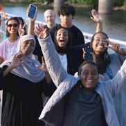 A group of smiling people of diverse race, ethnicity, gender and age, walk together. Some wear headscarves.