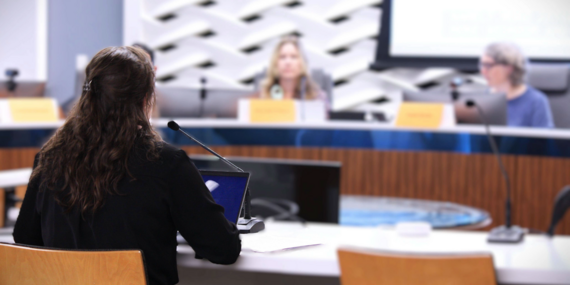 A woman in the foreground faces a panel of people in a meeting for the Planning Commission