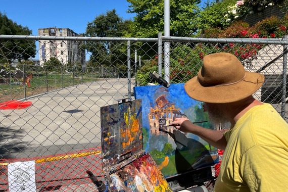 A person paints on a canvas the scene in front of him: Fields Park and Centennial Mills building with an explosion and collapsing water tower.