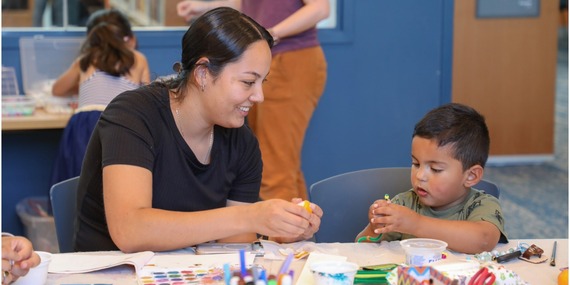 An adult smiles while working with a child on arts and crafts