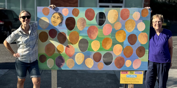 ARTS - Two people standing on either side of an art mural with one face drawn and shapes for other faces to be added.