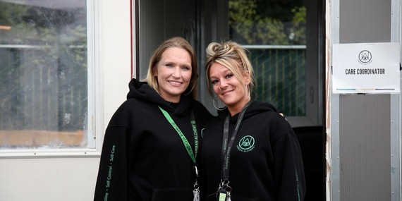 Two women wearing Urban Alchemy hoodies smile at the camera by a tiny house with a sign that says "care coordinator."