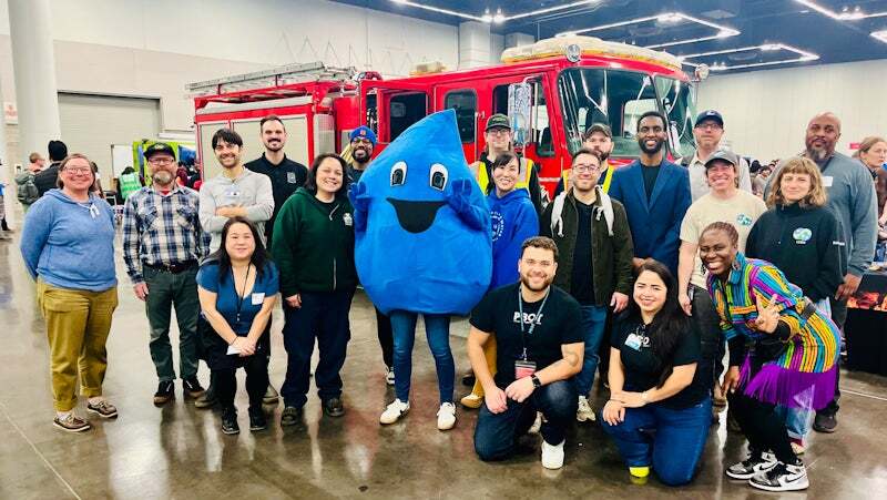 A group of smiling people, including city staff, pose with Drippy the Water Bureau mascot for WABT event (We Are Better Together)
