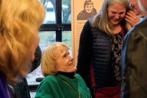 Jan Campbell, who uses a wheelchair, smiles with friends and supporters gathered near and a plaque with her photo in the background