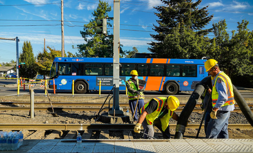 Three workers in bright safety vests perform maintenance along MAX tracks with a TriMet bus in the background