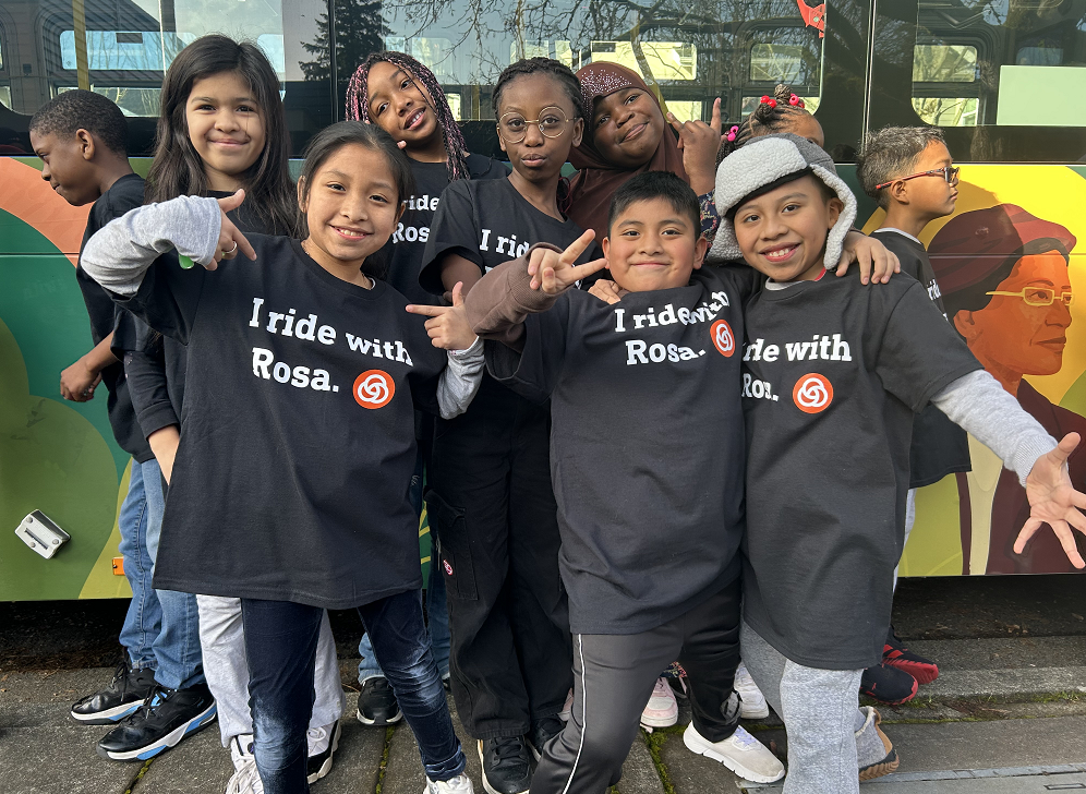 Image of Rosa Parks Elementary School students wearing "I Ride With Rosa" TriMet T-shirts in front of a commemorative TriMet bus