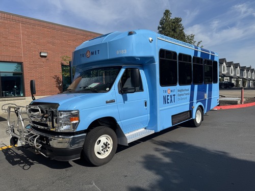 Image of a TriMet NEXT bus with a baby-blue color scheme, parked next to a brick building.