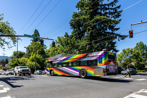 Image of TriMet's bus "From Progress to Beyond" on the road, with a cyclist in the foreground looking in its direction