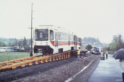 Train 103  being delivered, positioned on a temporary ramp structure. Workers in rain gear and bystanders with umbrellas observe the process