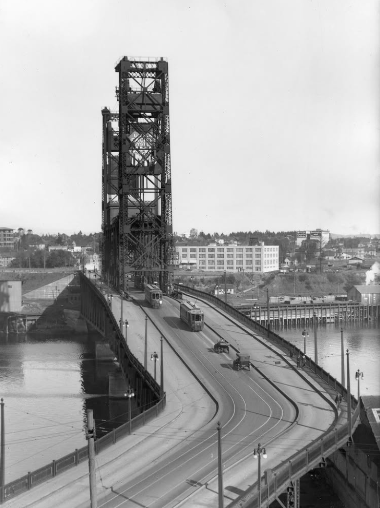 Black-and-white photo shows Portland’s Steel Bridge with streetcars and early automobiles crossing over the Willamette River.