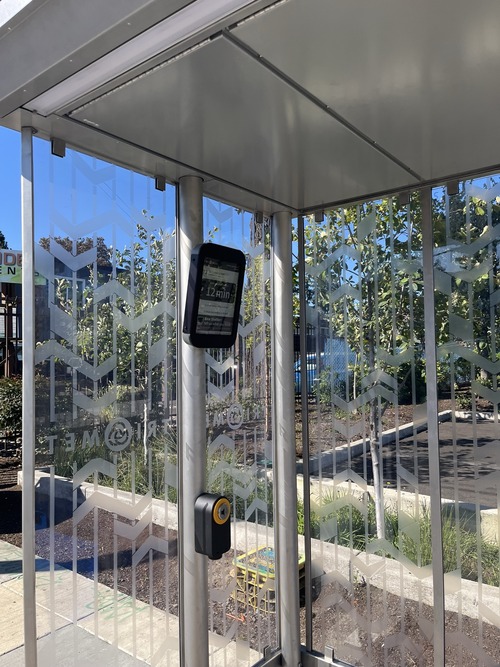A close-up inside a TriMet bus shelter, showing the electronic arrival display screen and a yellow push button on the support pole.