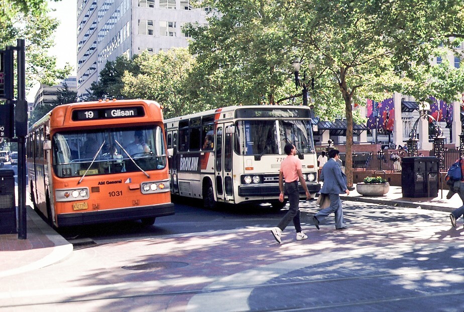 An orange bus and a white bus alongside it — with pedestrians crossing the street under the trees.