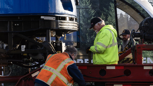 Three workers in high-visibility gear are working on equipment beneath the front end of a blue MAX train car that’s being offloaded