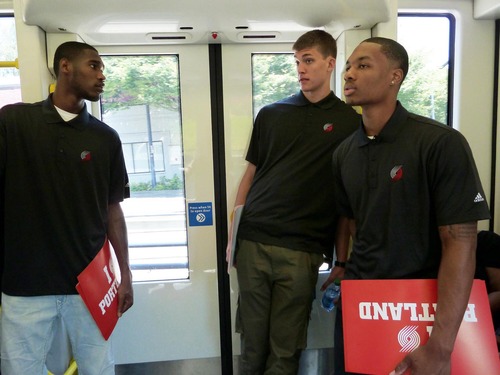 Three basketball players wearing matching black polo shirts with the Portland Trail Blazers logo are standing inside a MAX light rail train