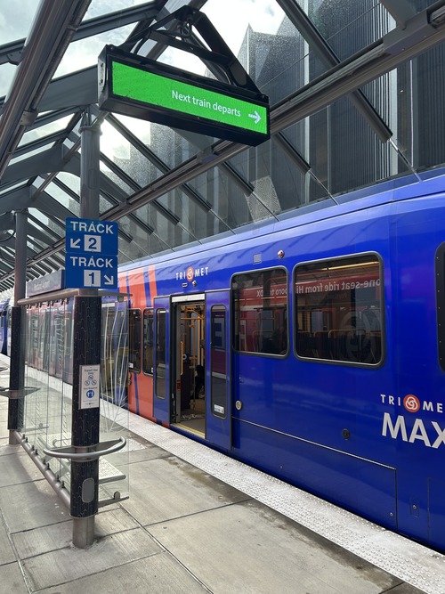 A MAX train sits at the PDX Airport station platform, with a digital sign overhead pointing riders toward the next departing train.