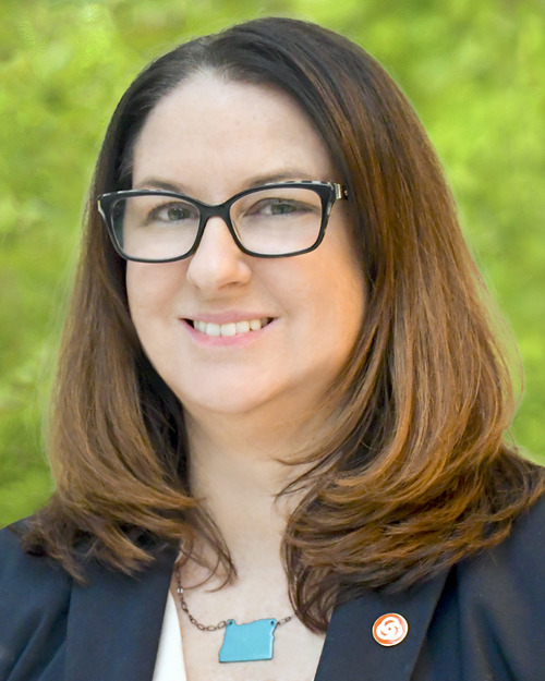 Jamie Snook, a woman with shoulder-length brown hair and glasses wearing a blazer with a TriMet pin and a necklace in the shape of Oregon
