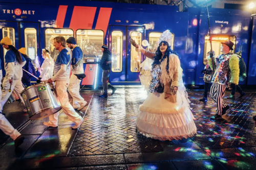 Costumed performers parade past a TriMet MAX train during a colorful nighttime festival.