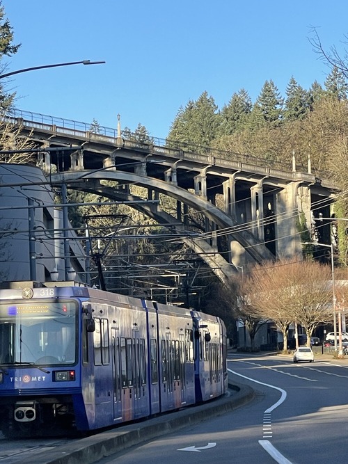 A MAX light rail train travels along a curved track beneath a large concrete arch bridge