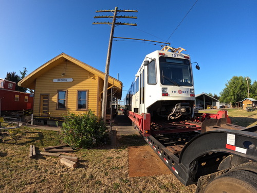 A light rail train sits on a trailer beside a small historic station building