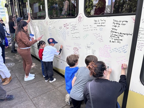 People of different ages stand beside a light rail train, writing messages and drawings across its exterior as others look on