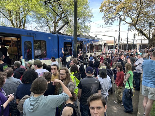 A large crowd of people stands around a MAX Type 1 train, many writing messages on its exterior while others watch and take photos