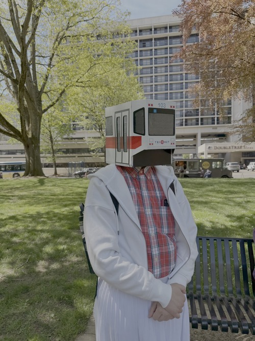 A person next to a park bench wearing a cardboard light rail train costume on their head, with trees and buildings in the background.