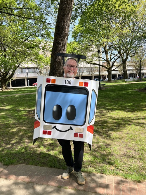 A smiling person stands in a park wearing a large cardboard light rail train costume with a face design on the front.