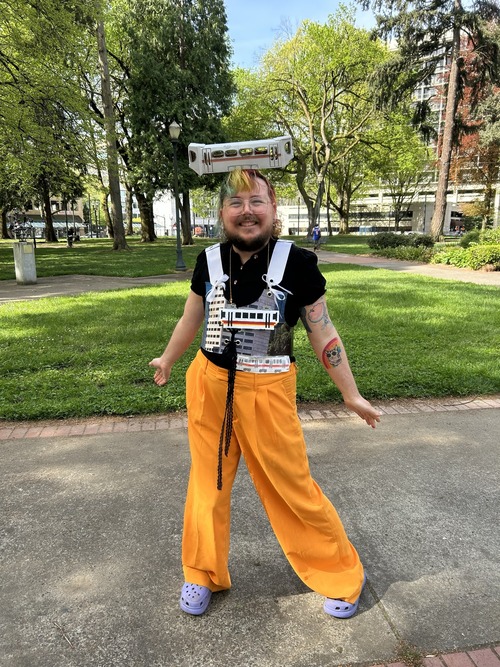 A smiling person stands in a park wearing a colorful outfit with handmade light rail train pieces attached, including one balanced on their head