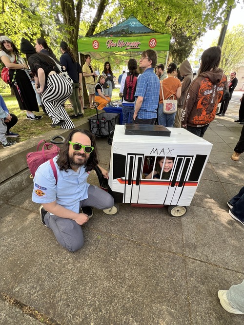A person kneels beside a child sitting inside a small, handmade MAX train, while a crowd gathers behind them