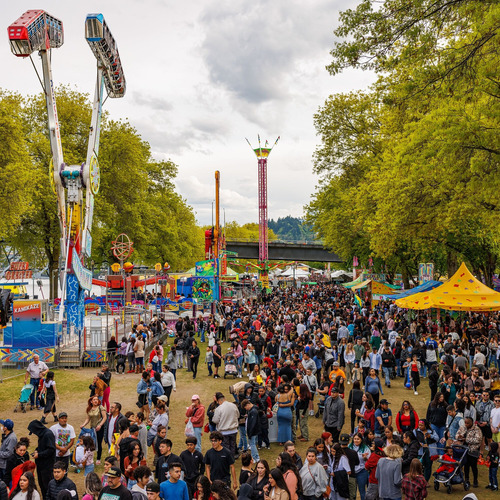 A large crowd fills a park at a lively fair, with colorful rides, game booths and tents lining the walkway under tall trees