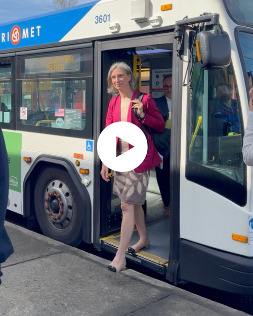 Maxine Dexter steps off a TriMet bus onto the sidewalk, with another passenger visible behind her and trees in the background.