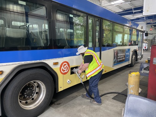 A TriMet maintenance worker in a safety vest and wide-brimmed hat fuels a bus