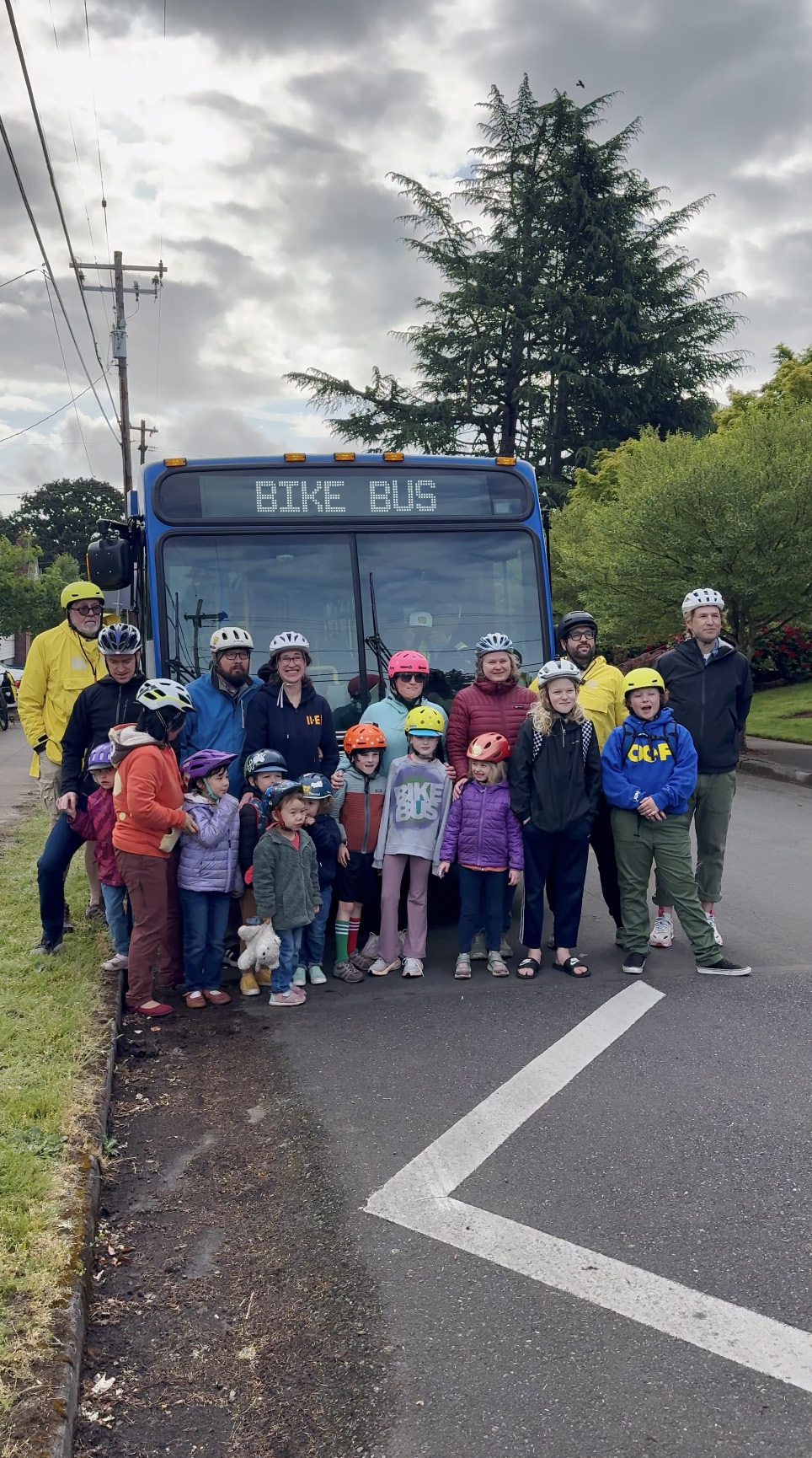 Group of adults and children wearing bike helmets gathered in front of a TriMet bus displaying “BIKE BUS.”